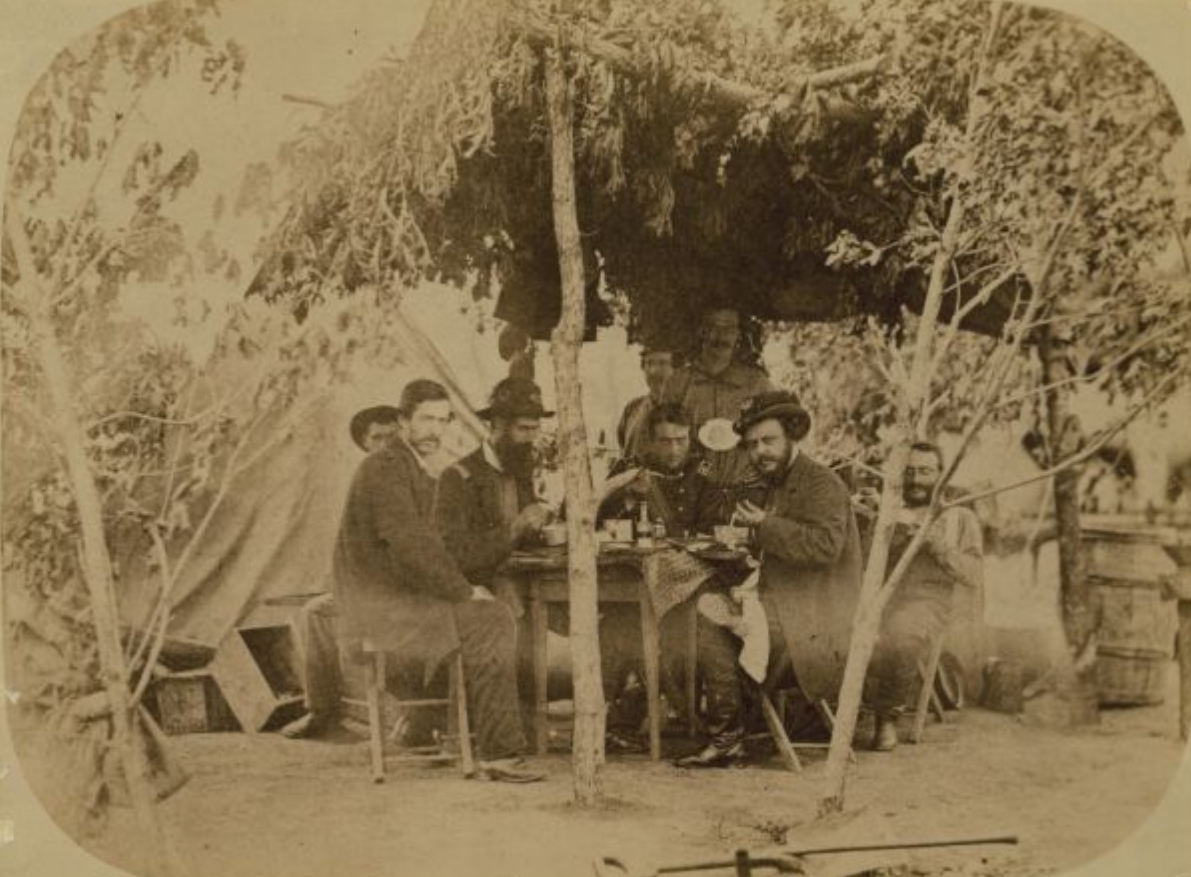 A group of Soldiers sit around a table under a tarp held up by trees and branches. 