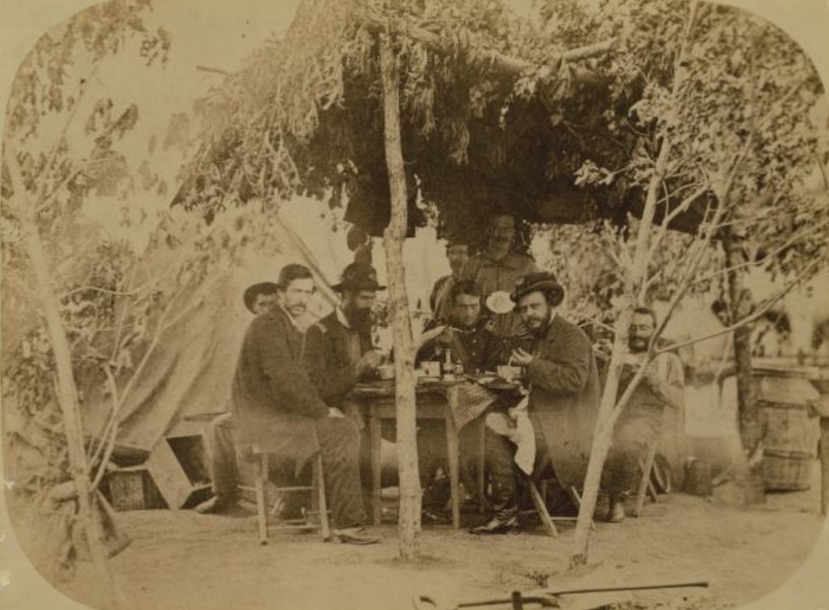A group of Soldiers sit around a table under a tarp held up by trees and branches.