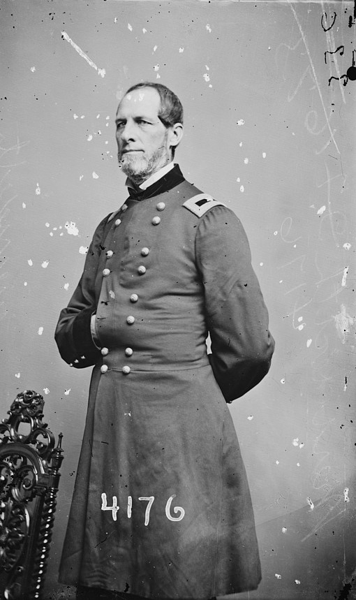 Man standing next to an ornate chair wearing a Civil War uniform jacket. His right hand is in his jacket, left hand behind his back, looking to the left of the camera.