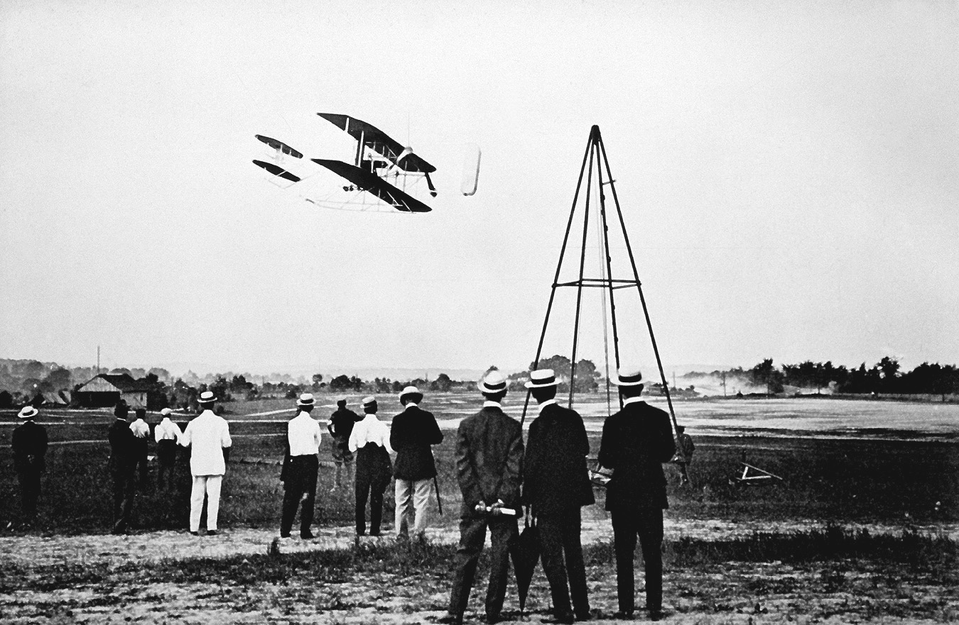 Men stand looking up at the Wright Flyer in the air. 