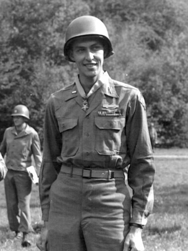 Man stands in a field in a World War II era Army field uniform wearing a helment and a Medal of Honor around his neck. 