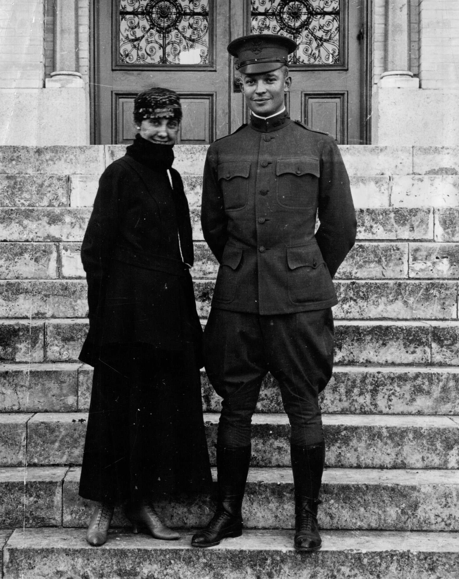 Woman in a long dress and coat stands next to a man in a World War I era Army uniform. They stand on stone steps in front of an ornate door.