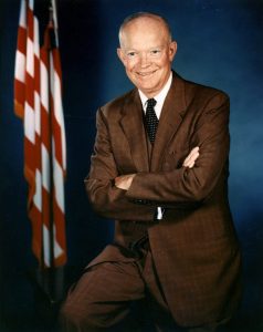 Eisenhower wearing a brown suit leans against a desk, crossing arms and smiling at the camera, with an American flag in the background.