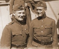 Two Soldiers in World War I uniforms stand side-by-side smiling. 