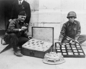 Two Soldiers kneel next to a box of recovered objects. The man on the left holds up an small object in his right hand for the camera to see. 