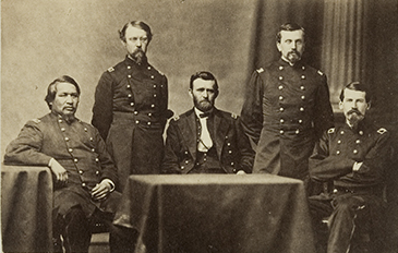Three Soldiers in Civil War era uniforms sit behind a table with two Soldiers standing behind. 
