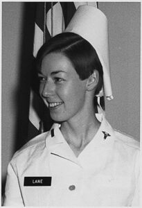 Woman in a nurses uniform standing in front of an American flag looking to her right. 