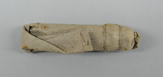 Paper cartridge consisting of folded over tube of gray laid paper containing black powder, lead ball and buckshot all secured with a linen string. Object ID 2013.0326. Courtesy of The Fort Ticonderoga Museum Collection.