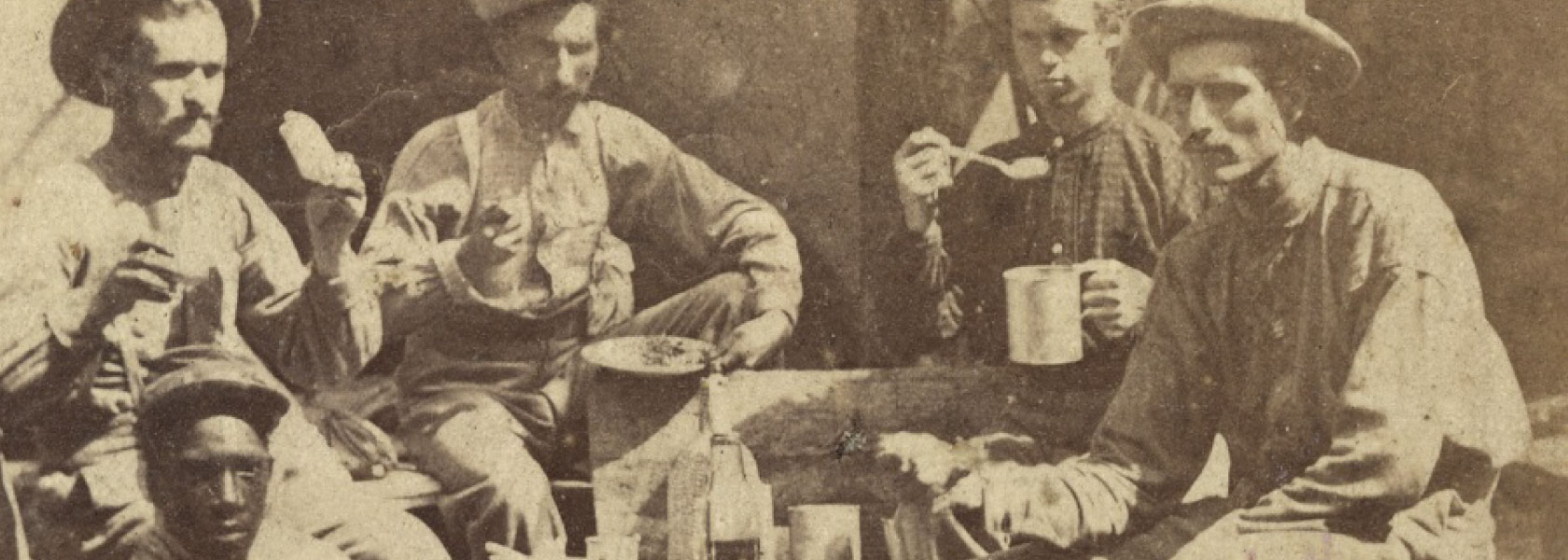Black-and-white historical photograph of four men sitting together outdoors, eating and drinking from plates, cups, and a large container.